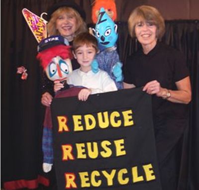 Image of Peppermint puppetry and the three Rs, perfect together—puppeteers Lois and Caroll, along with a student who has just observed one of their programs, pose with three of their puppets (one is made from a plastic bottle) and butterfly scenery made from a hanger and a Mylar balloon